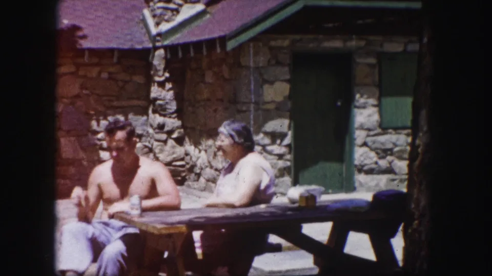 Two adults converse or pause at a picnic table in front of a rustic stone cabin in Colorado.