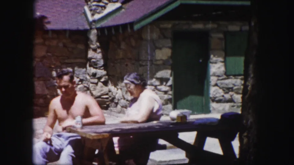 The 1958 Colorado scene holds on two adults resting at a wooden table outside a stone cabin.