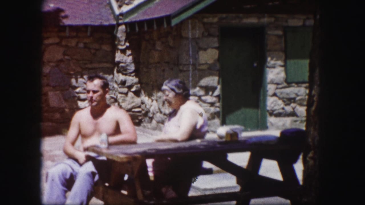 In 1958 Colorado home-movie footage, a shirtless man and older woman sit at a wooden picnic table near a rustic stone cabin.