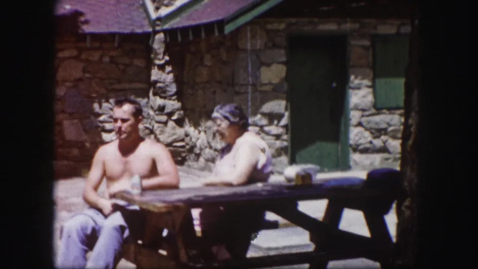 In 1958 Colorado home-movie footage, a shirtless man and older woman sit at a wooden picnic table near a rustic stone cabin.