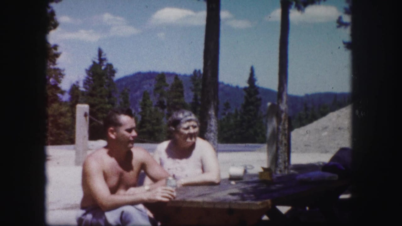 A shirtless man and an older woman sit outdoors at a picnic table in Colorado, with pines and distant mountains visible behind them.