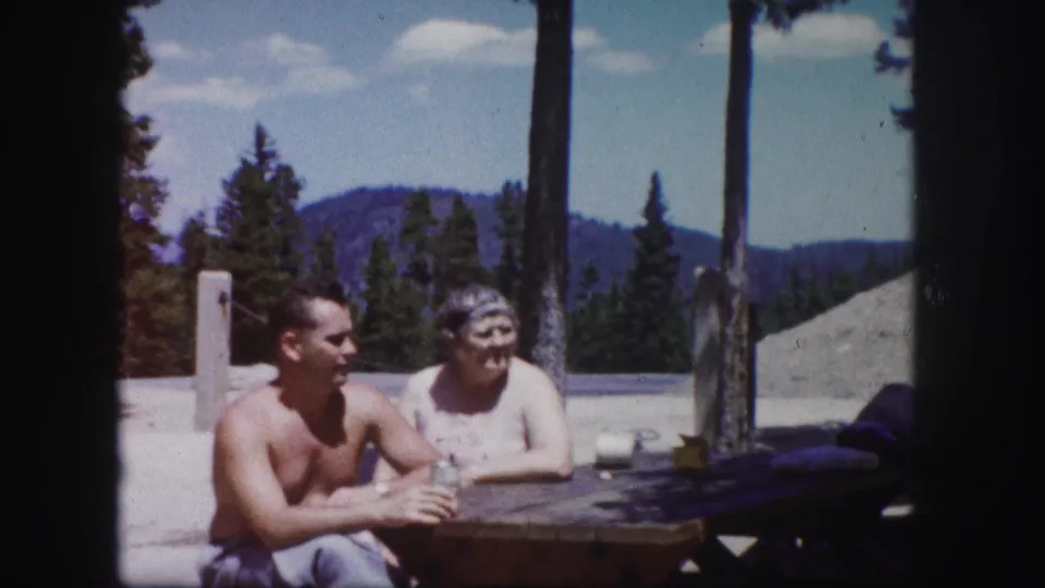 A shirtless man and an older woman sit outdoors at a picnic table in Colorado, with pines and distant mountains visible behind them.