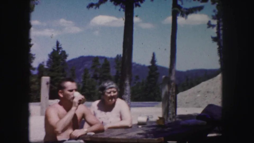 The man raises a drink at a picnic table while the older woman sits beside him in the 1958 Colorado mountain setting.
