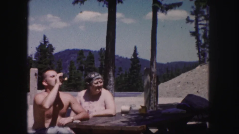 A shirtless man lifts a drink beside an older woman at an outdoor picnic table, with Colorado pines and mountains in the background.