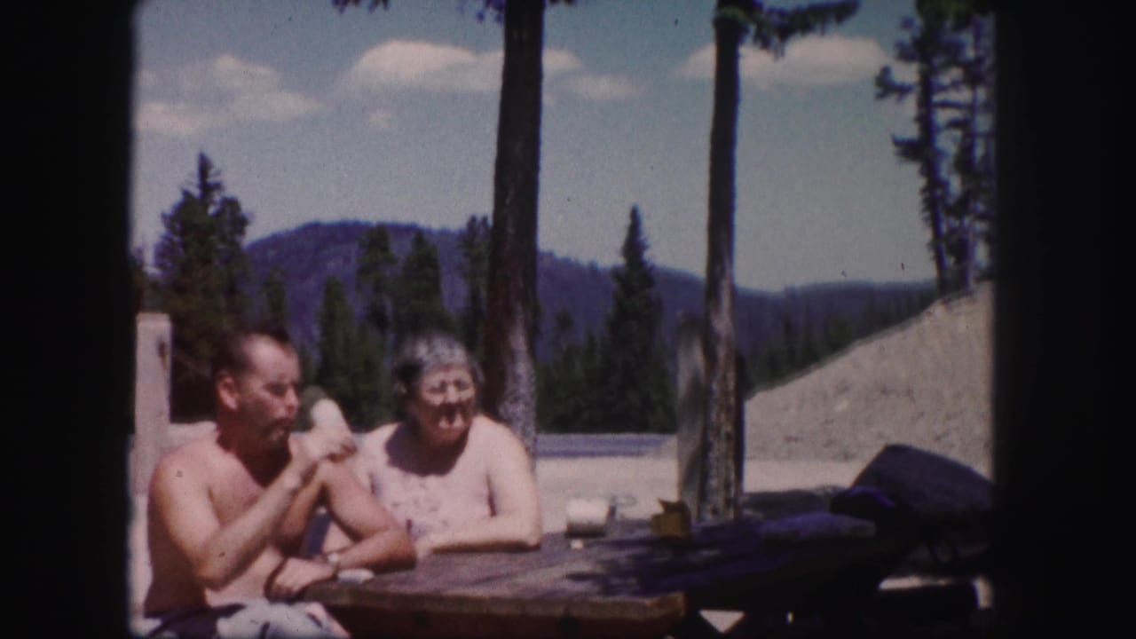 In 1958 Colorado home-movie footage, a shirtless man and woman sit at a picnic table against trees and distant mountains.