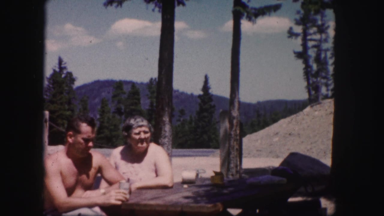 The couple remains seated at a wooden table in Colorado, with tall trees, mountains, and a bright sky visible behind them.