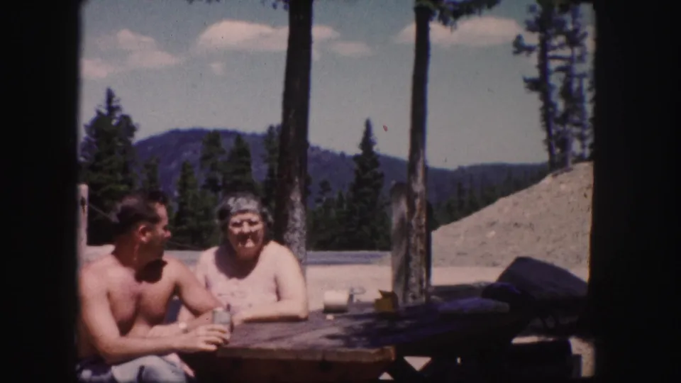 A candid 1958 Colorado frame shows the man turned toward the woman while they sit at a picnic table outdoors.