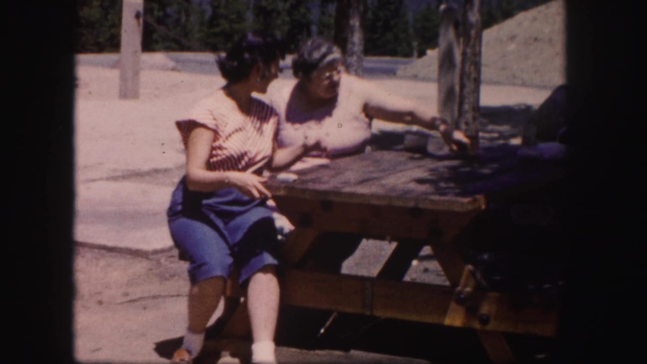 In 1958 Colorado home-movie footage, two adult women sit beside a wooden picnic table in bright sun.