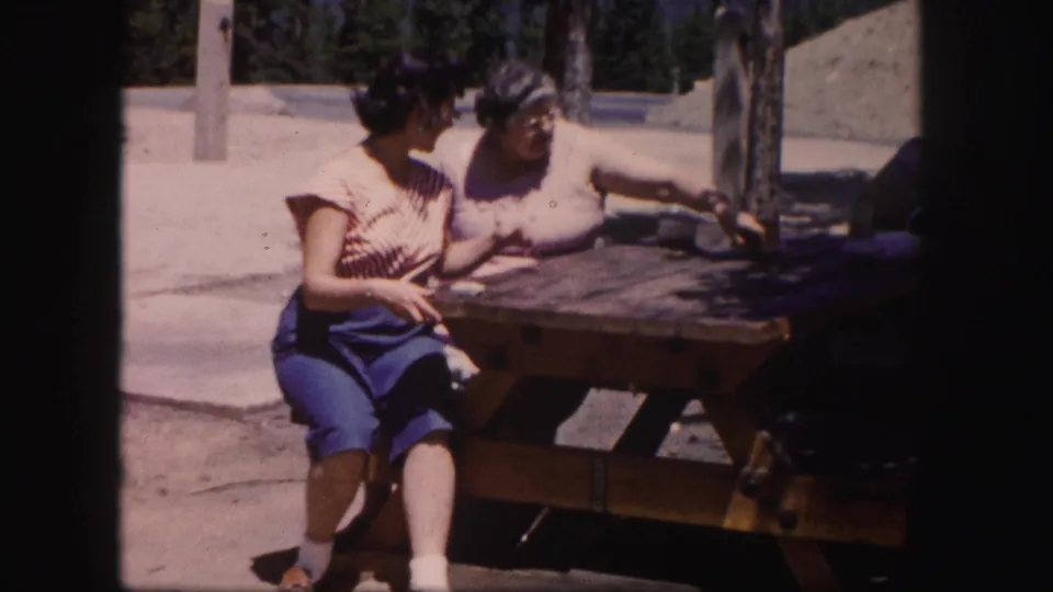 In 1958 Colorado home-movie footage, two adult women sit beside a wooden picnic table in bright sun.