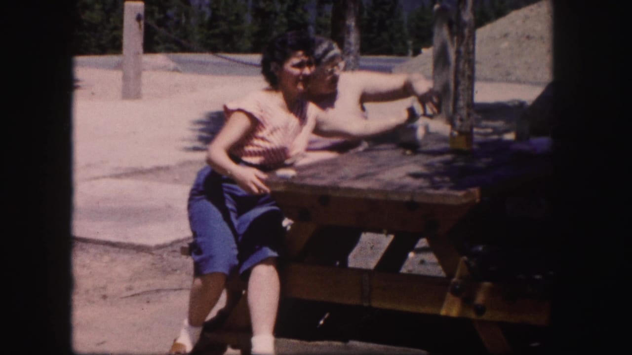 Two adult women at a wooden picnic table lean and gesture during a sunny 1958 Colorado home-movie scene.