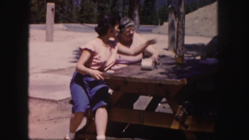 A 1958 Colorado frame shows two women seated close together while one gestures across a wooden picnic table.