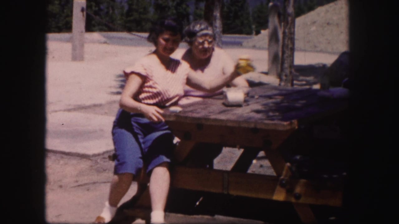 In 1958 Colorado home-movie footage, two women sit at a wooden picnic table in bright sun with trees and pavement behind them.