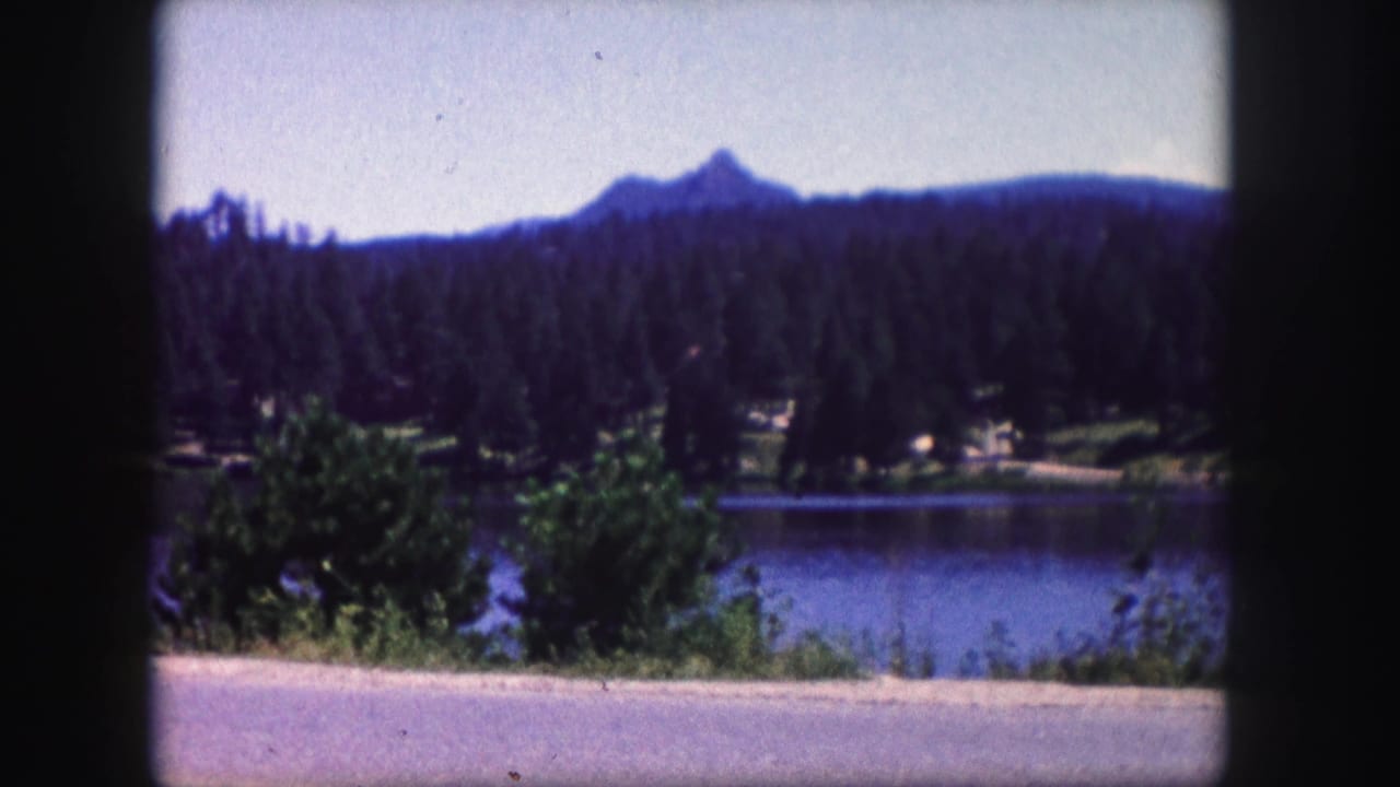 Blue lake below evergreen hills with a distant mountain peak and roadside foreground.