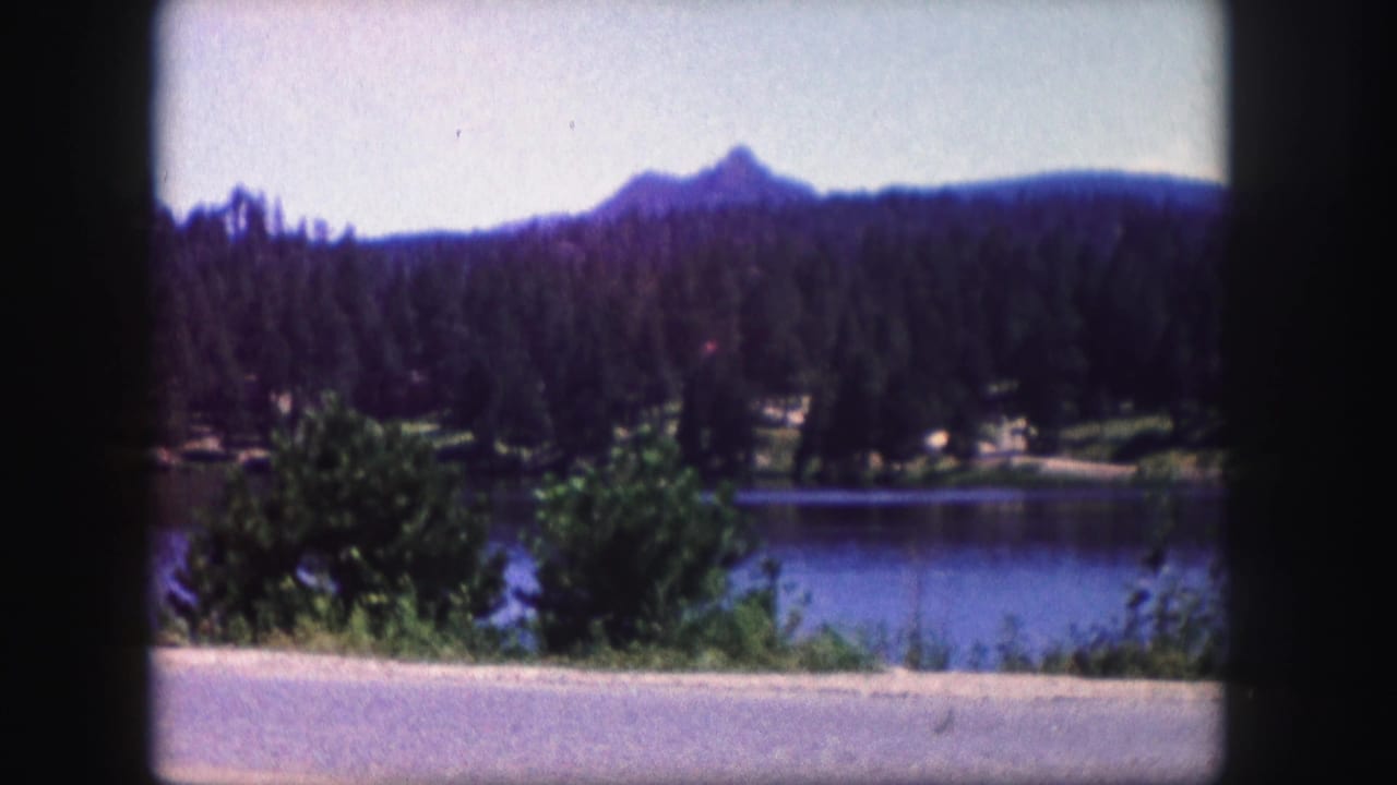 Colorado home-movie landscape with evergreen hills, calm blue water, roadside foreground, and a distant mountain peak under a pale sky.