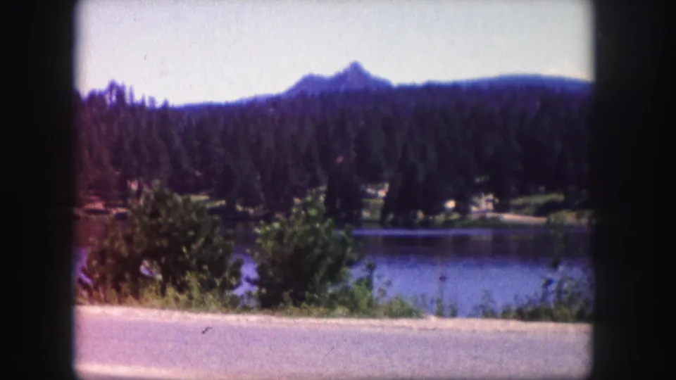 Vintage 8mm Colorado frame with a lake in the middle ground, dark evergreen hills, foreground shrubs, and a pointed mountain peak in the distance.