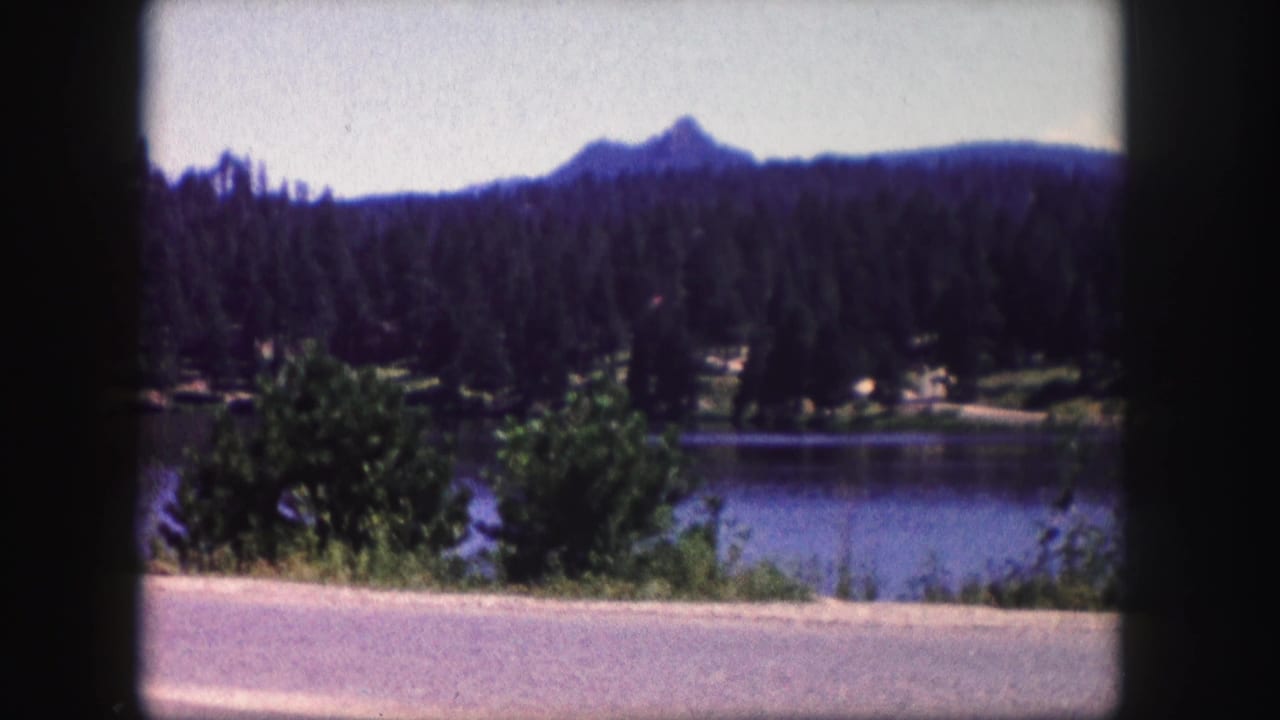 1958 Colorado 8mm view of a lake bordered by pine forest, with a distant mountain peak and road in the foreground.