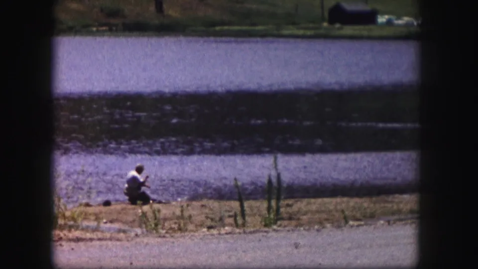 Distant 1958 Colorado lakeside frame showing a person crouched near reeds along the water's edge.