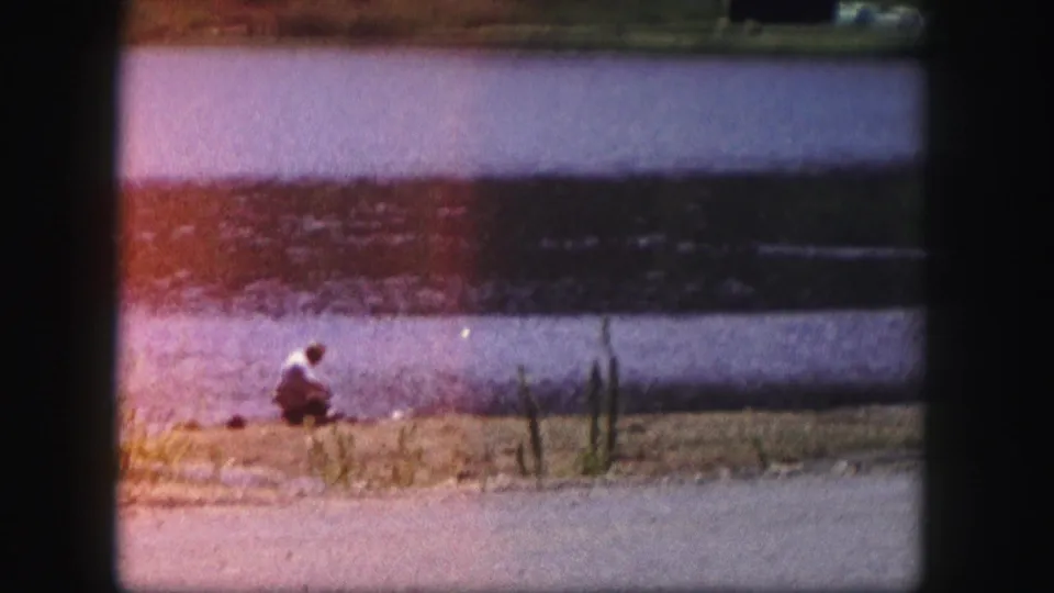 1958 Colorado home-movie frame of a person crouched by a lake shore, with visible film color shift and light leak.