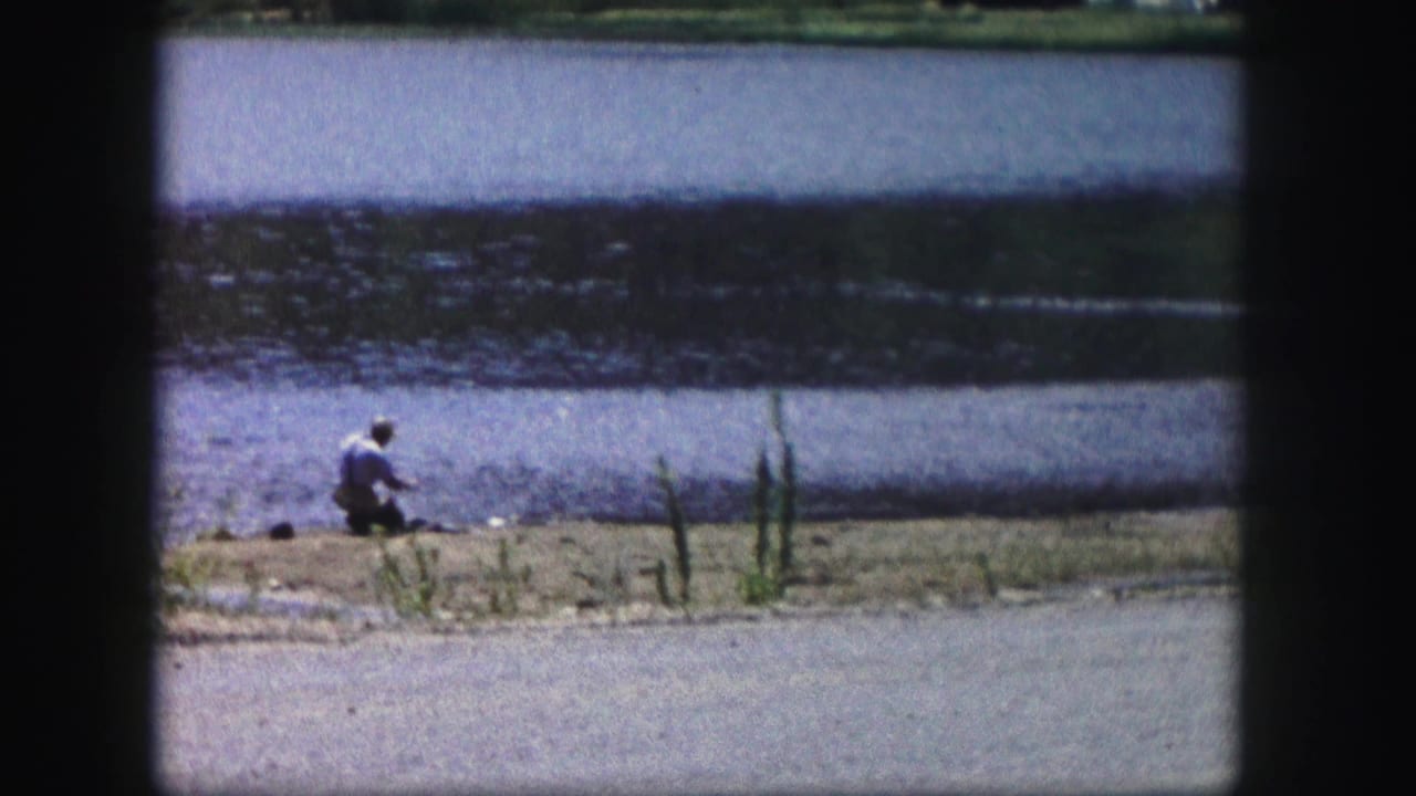 1958 Colorado 8mm frame showing a lone man in a light shirt fishing beside calm lake water.