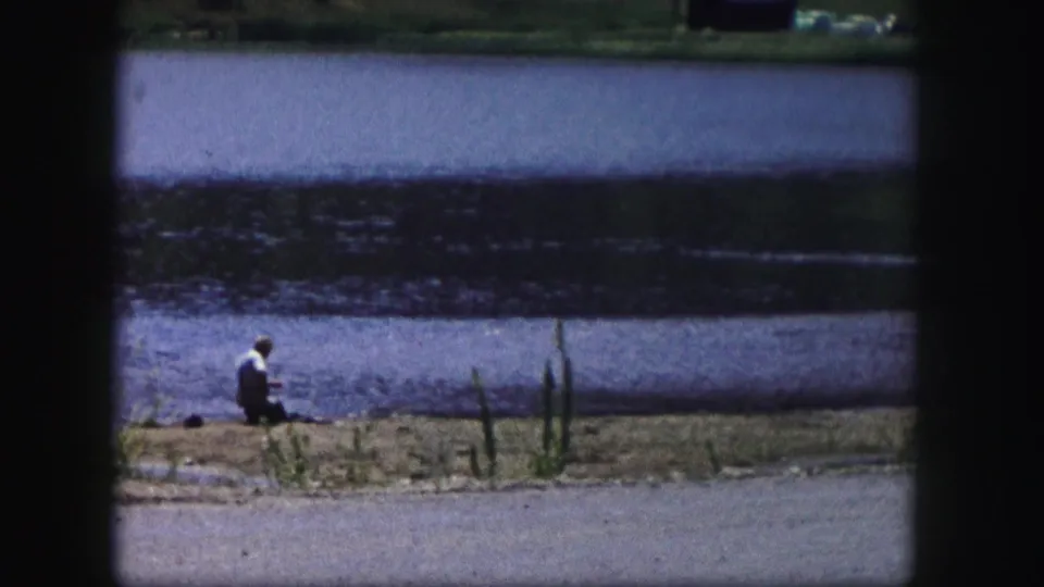 A lone adult fisherman sits at the edge of a Colorado lake in this 1958 home-movie frame.