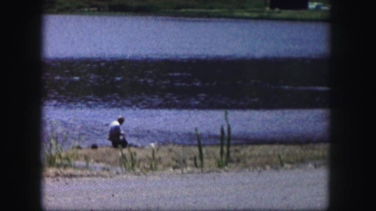 1958 Colorado home-movie frame showing a lone person near a lake or pond, with reeds, shoreline, and road visible in soft 8mm color.