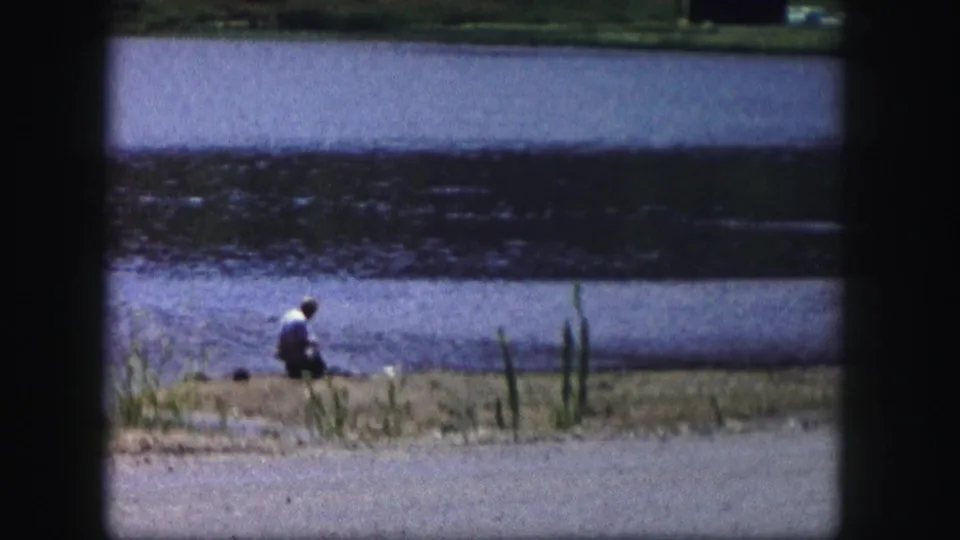 1958 Colorado home-movie frame showing a lone person near a lake or pond, with reeds, shoreline, and road visible in soft 8mm color.