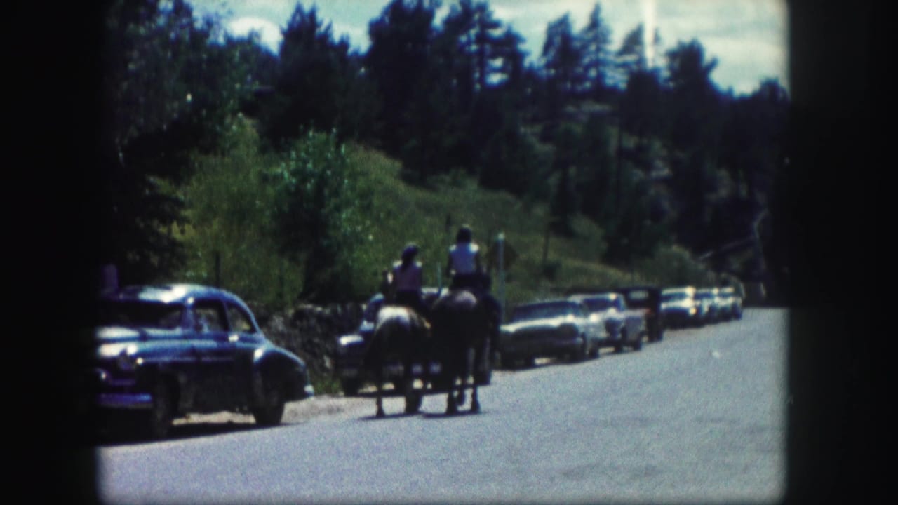 Two horseback riders move along a Colorado roadside in 1958, passing parked mid-century cars with trees and low hills behind them.