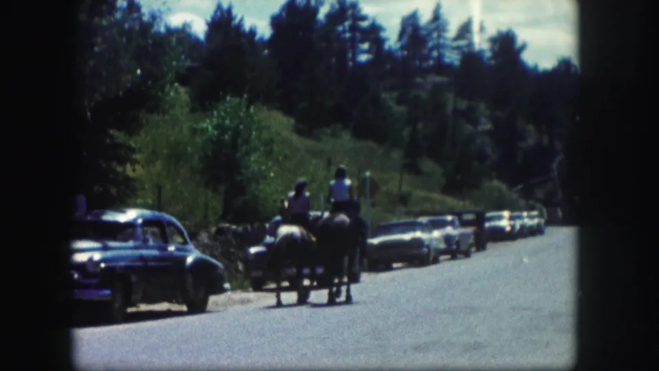 Two horseback riders move along a Colorado roadside in 1958, passing parked mid-century cars with trees and low hills behind them.