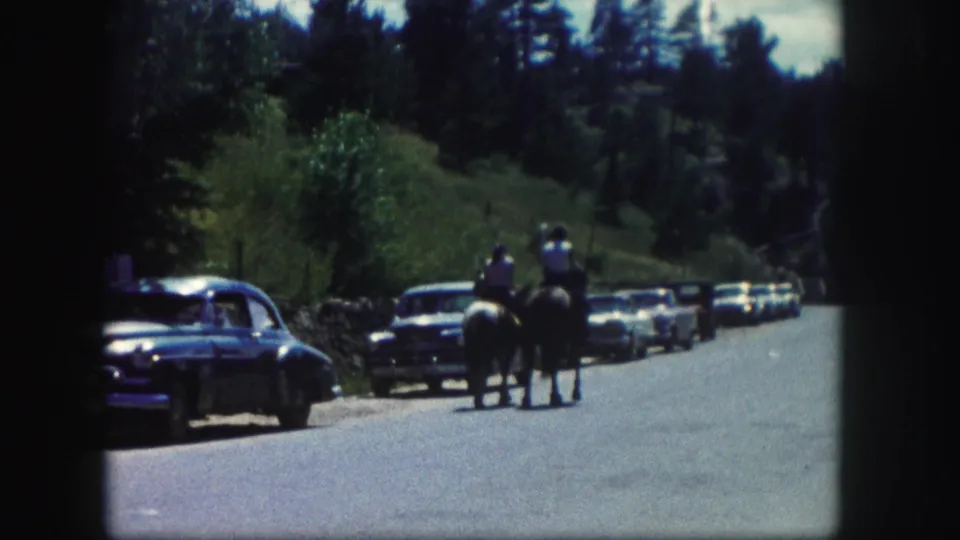 1958 Colorado roadside view of two people riding horses away from the camera, with parked cars, trees, and hillside in the background.
