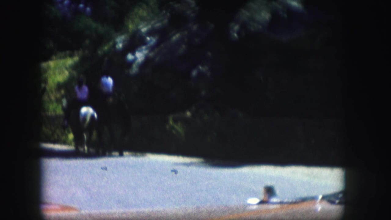 In 1958 Colorado home-movie footage, two distant horseback riders travel along a curving paved road lined with dense greenery.