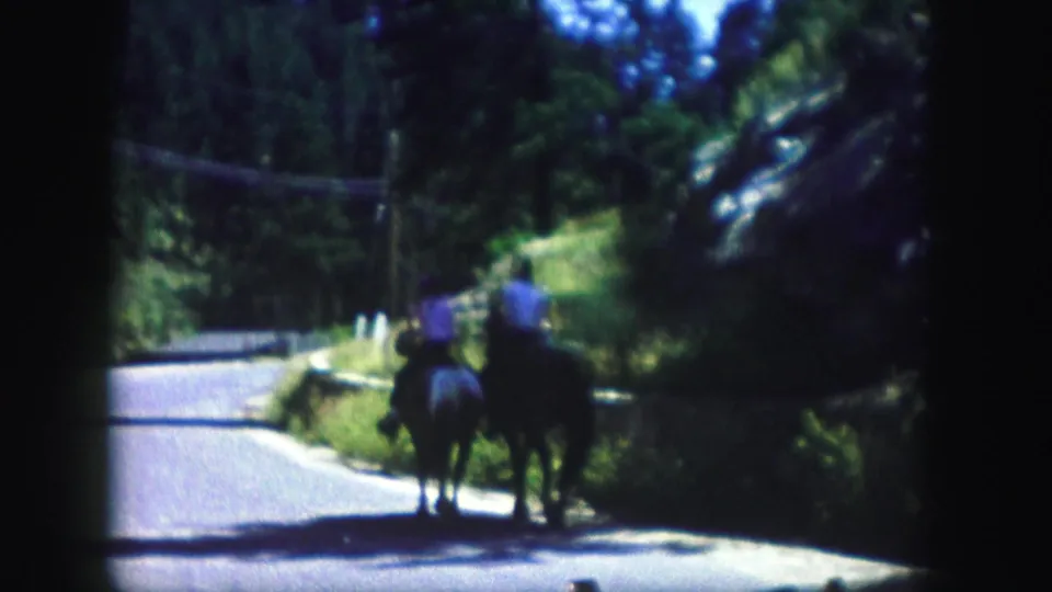 Two horseback riders appear on a winding Colorado road, with trees, hillside vegetation, and overhead lines in the background.