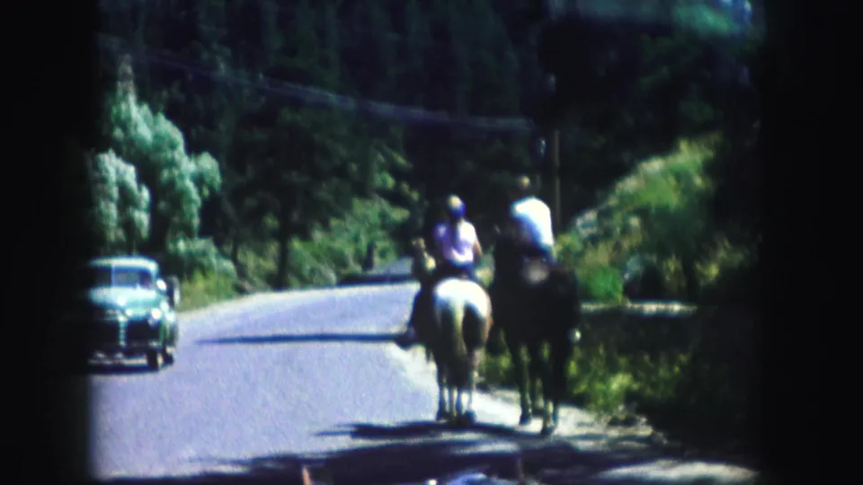 In the wooded Colorado road scene, two riders approach the camera while a midcentury car is visible along the opposite side.