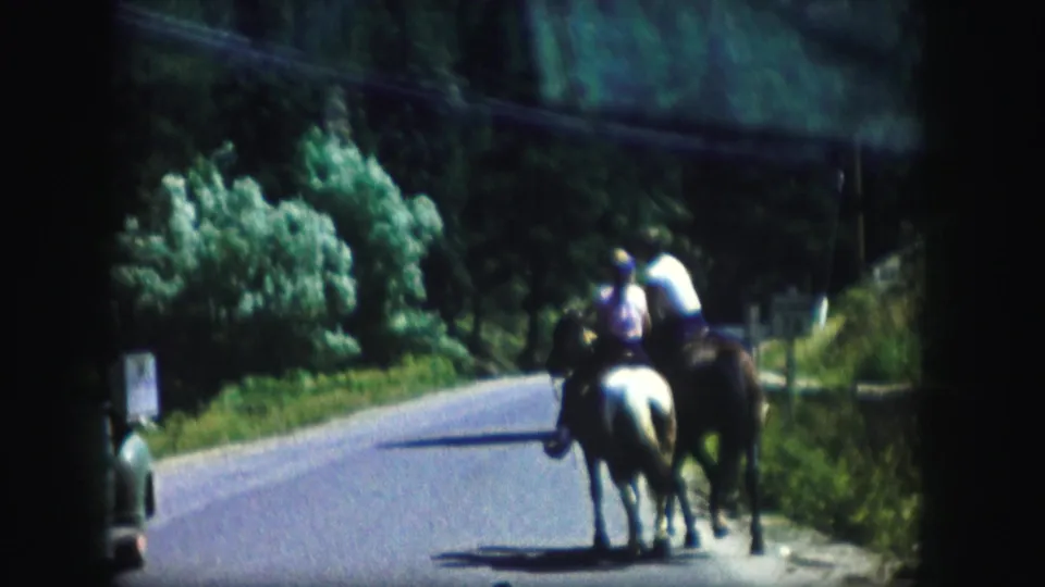 Two horseback riders, one on a light horse and one on a dark horse, pass along the edge of a wooded Colorado road in 1958 footage.