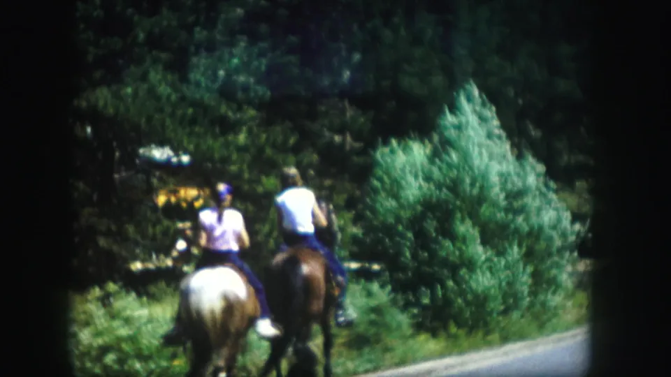 Two horseback riders, one on a light horse and one on a darker horse, continue along a rural Colorado road.