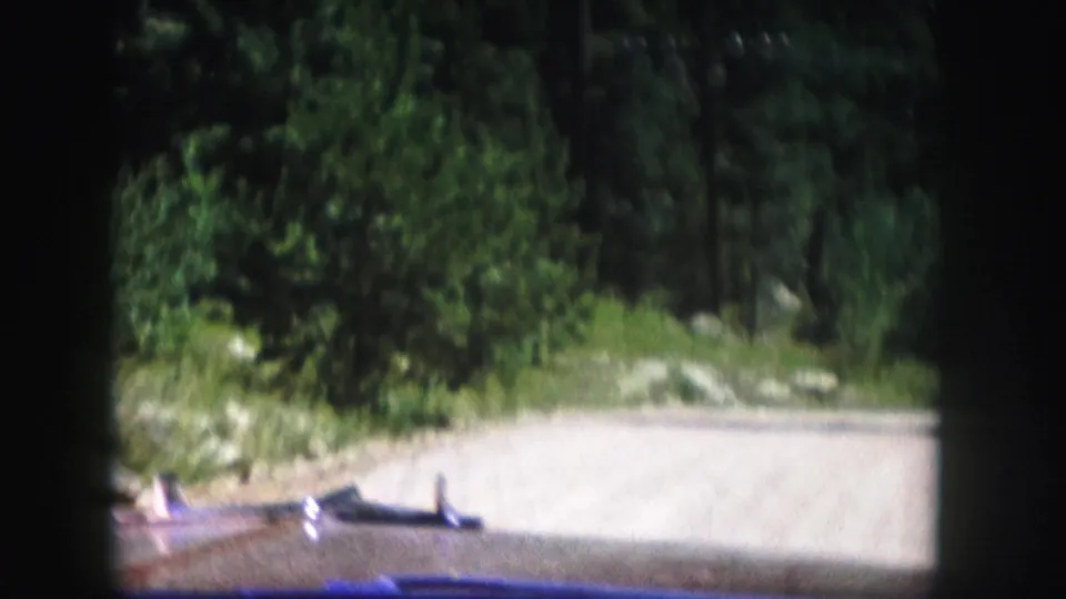 The road continues through a wooded Colorado setting, with trees and rocks visible at the roadside in the 1958 footage.