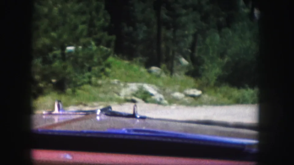 1958 Colorado 8mm frame showing a car hood and chrome trim facing a pale forest road lined with trees and rocks.