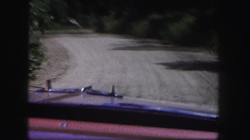 8mm home-movie frame from Colorado showing the car’s front edge below a forest road with trees and roadside shadow.