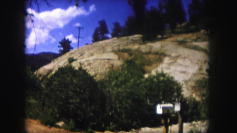 1958 Colorado roadside view with a white mailbox, shrubs, rocky hillside, utility poles, and blue sky.