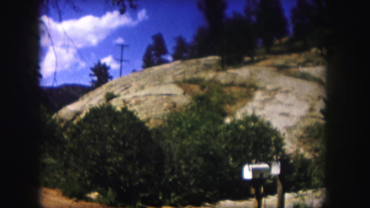 1958 Colorado 8mm frame showing a white mailbox, green shrubs, rocky hillside, utility pole, trees, and blue sky.