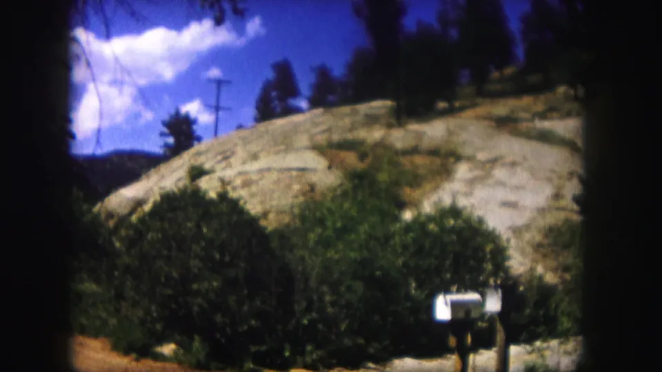 1958 Colorado 8mm frame showing a white mailbox, green shrubs, rocky hillside, utility pole, trees, and blue sky.