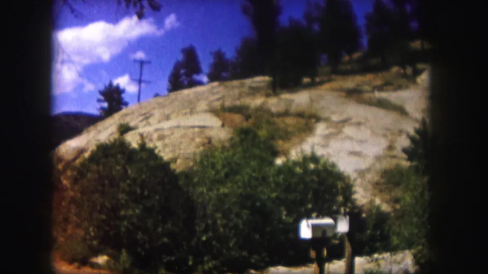 1958 Colorado 8mm view of a mailbox and shrubs below a sunlit rocky hillside with trees and clouds overhead.