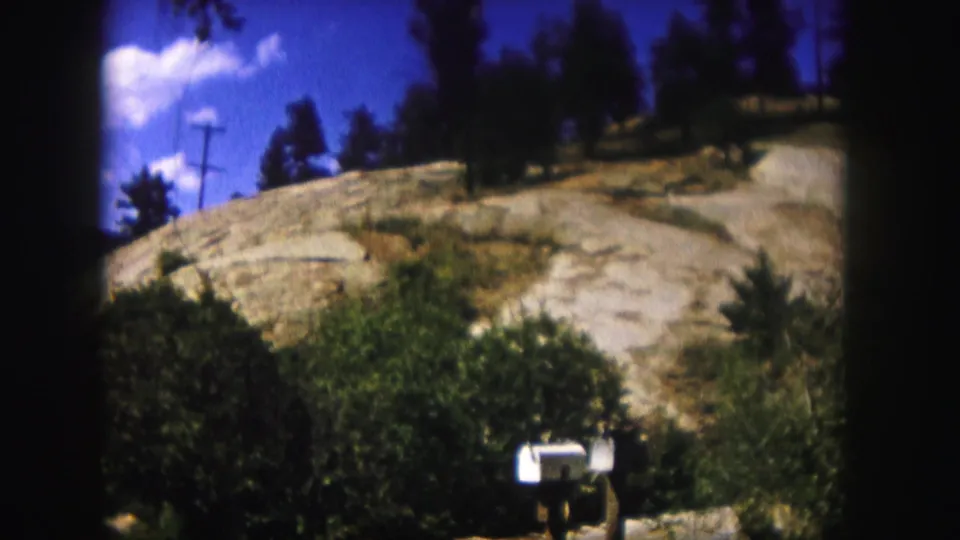 1958 Colorado frame showing a white mailbox at the base of bushes and a rocky hillside beneath a blue sky.