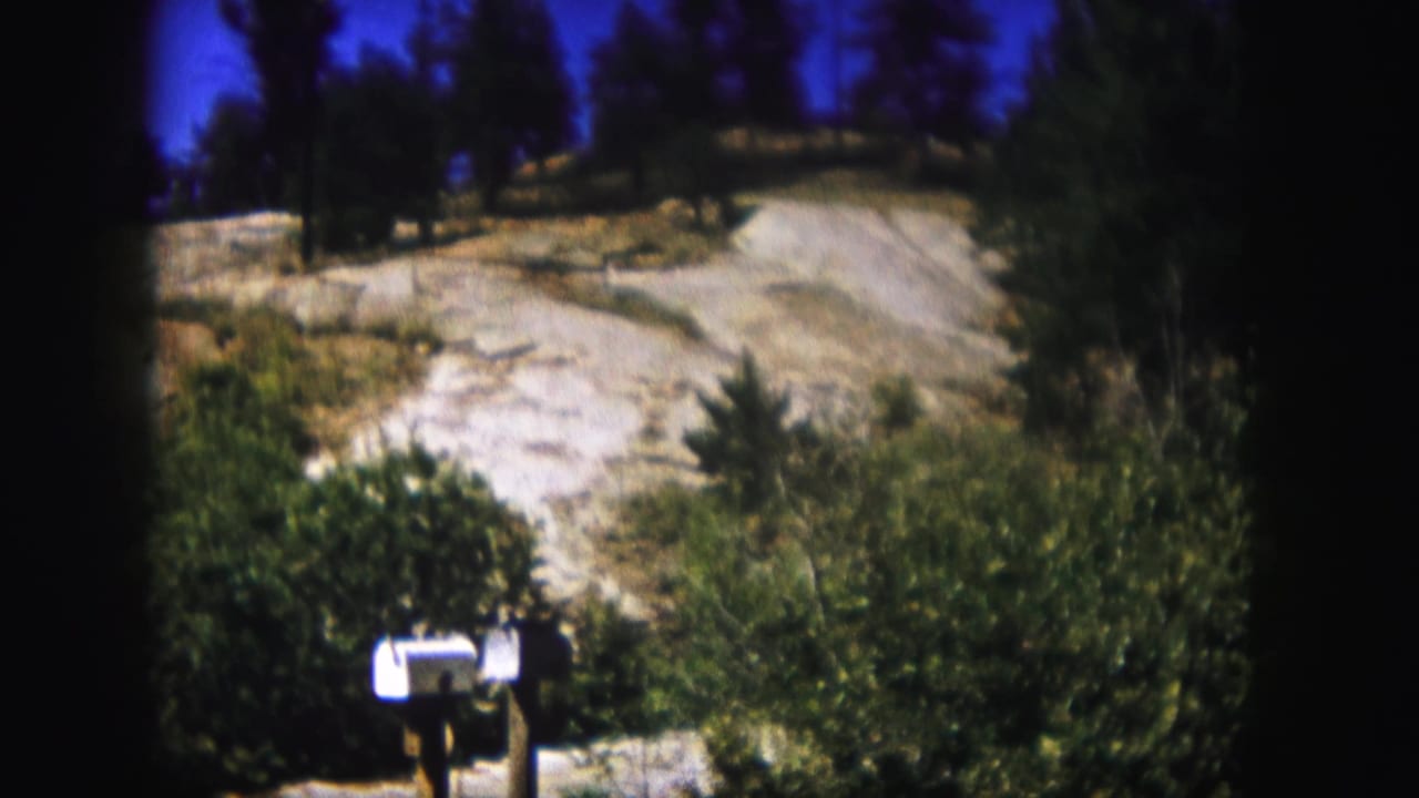 1958 Colorado home-movie frame showing a white roadside mailbox below shrubs, a pale road or driveway, dry hillside, trees, and deep blue sky.