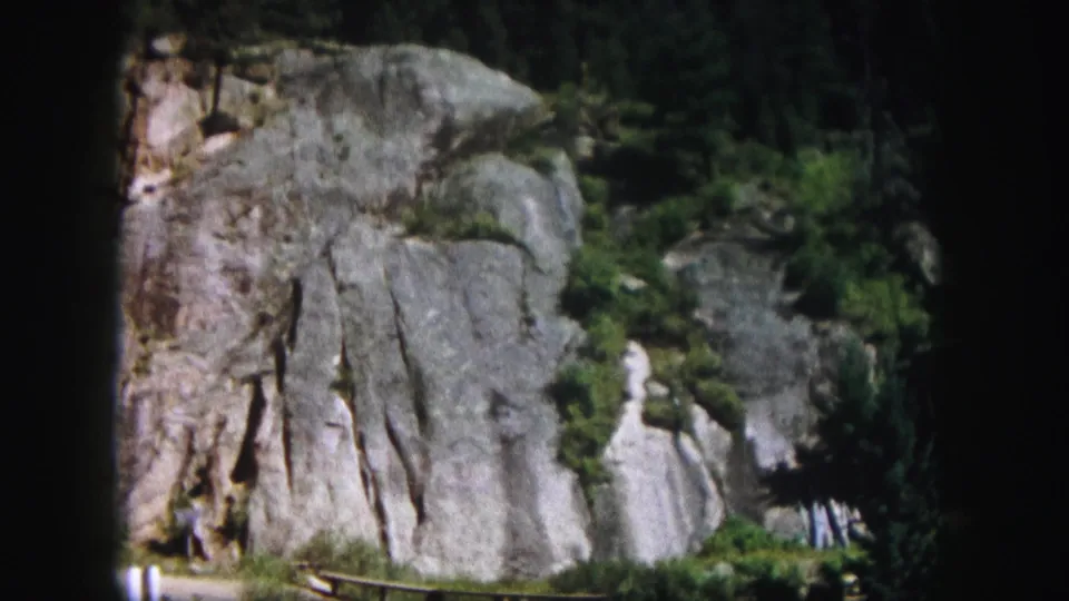 1958 Colorado landscape frame showing a textured rock face with vertical striations, scattered greenery, and trees near the base and edges.