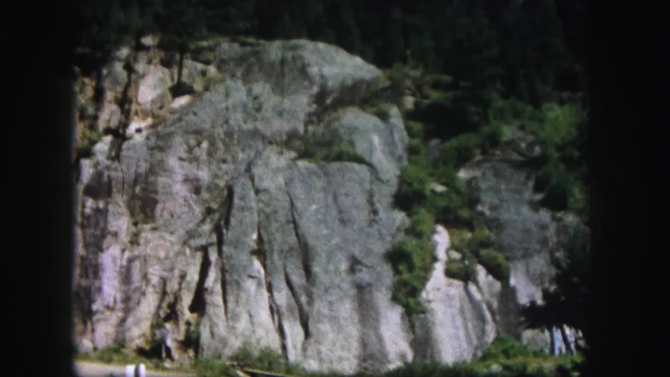 1958 Colorado home-movie view of a gray rocky cliffside with vertical fissures, patches of greenery, and pine trees along the lower edge.