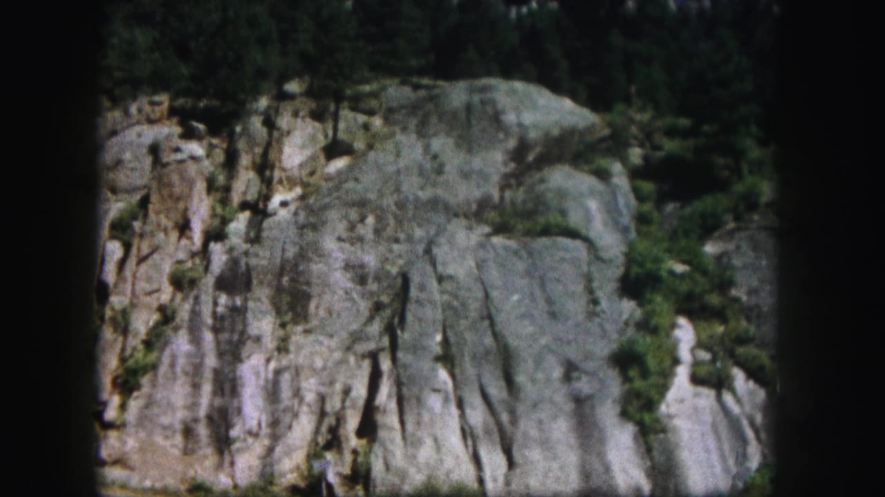 1958 Colorado mountain view with pale stone, deep cracks, scattered greenery, and a dark 8mm frame edge.