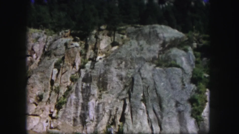 1958 Colorado 8mm frame centered on fissured roadside rock with pine growth and visible film-edge shadowing.