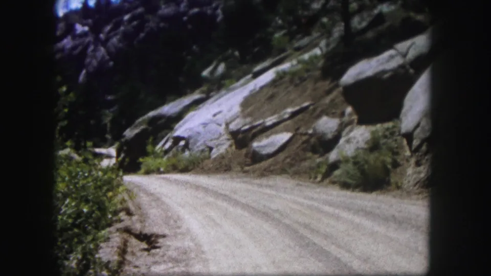 1958 Colorado 8mm view of a pale dirt road curving through rock cuts with forested slopes nearby.