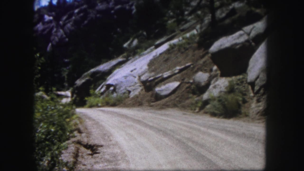 1958 Colorado 8mm frame showing a pale dirt road beside large boulders, brush, and evergreen trees under bright sky.