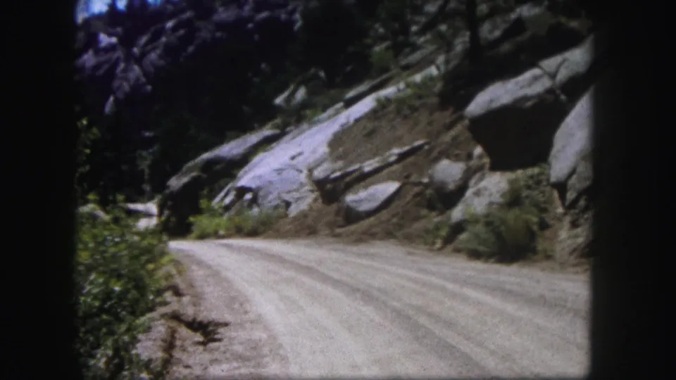 1958 Colorado 8mm frame showing a pale dirt road beside large boulders, brush, and evergreen trees under bright sky.